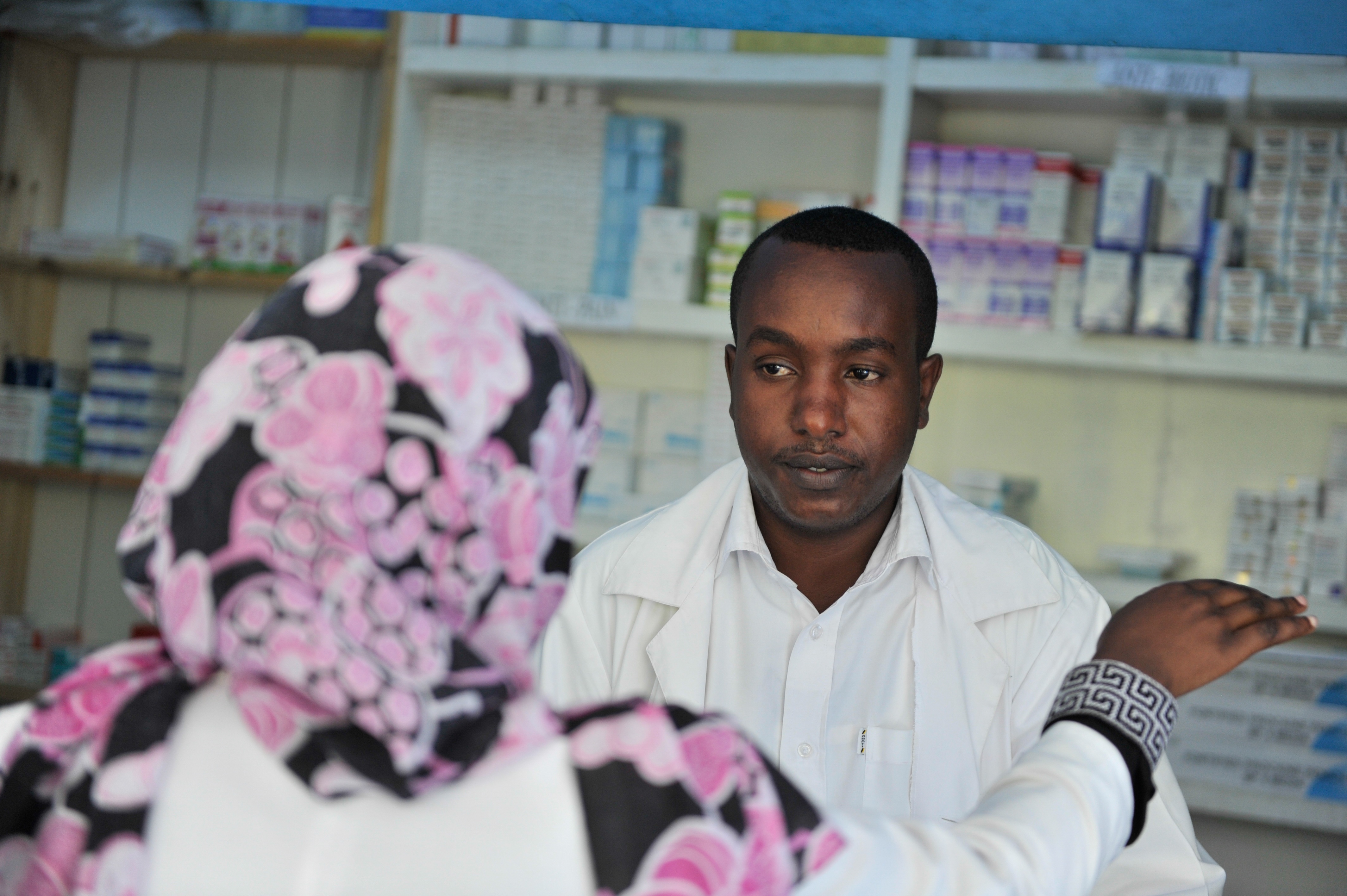 Eine Apothekerin in einem weißen Kittel spricht mit einer sitzenden Frau in einer Apotheke mit Regalen voller Schachteln und anderen Gegenständen dahinter.
