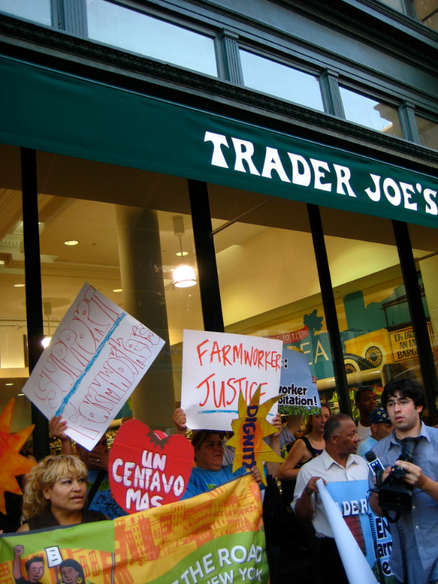 Eine Gruppe von Menschen steht in der Nähe eines Ladens und hält Schilder und Spielkarten, während im Hintergrund eine Säule und Dachleuchten zu sehen sind.