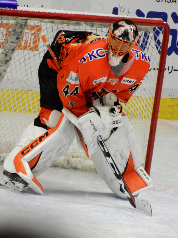 Ein Hockey-Torwart in einem orange-weißen Dress mit Helm und Handschuhen hält vor einem Tor, einen Stock in der Hand, mit sichtbarem Torpfosten und einer Tafel im Hintergrund.