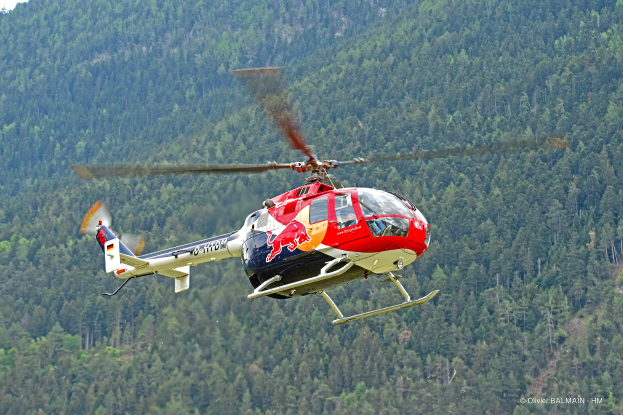 Roter Bullenhubschrauber über einem grünen Wald mit Hügeln und einem klaren blauen Himmel fliegt.