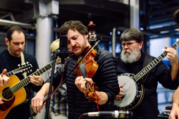 Eine Gruppe von Musikern in schwarzer Kleidung, die Instrumente spielen, mit einem Geigenspieler vor einem Mikrofon und einem Gebäude im Hintergrund.