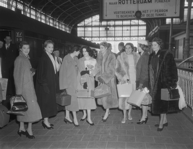 Black and white image of a group of women standing at a train station, each holding a bag, with a train on the left and railings and a board on the right.