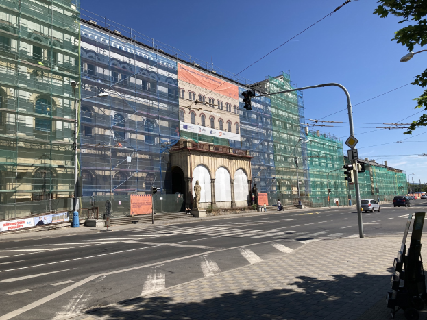 Großes modernes Gebäude im Bau in Berlin, umgeben von Straßeninfrastruktur, Fahrzeugen, Fußgängern und Bäumen mit bewölktem Himmel.
