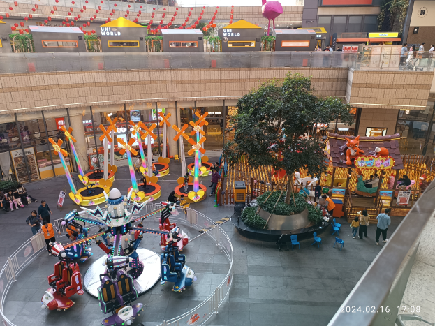 Ein Vergnügungspark innerhalb eines Einkaufszentrums mit verschiedenen Attraktionen, Bäumen, Menschen, Stühlen und Ständen, mit Gebäuden oben und einem Wasserzeichen unten rechts.