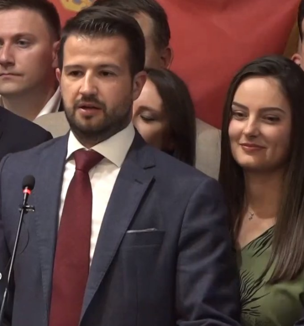 Ein Mann im Anzug spricht in ein Mikrofon an einem Rednerpult und wendet sich an eine Menge mit einem Banner im Hintergrund.