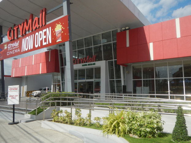 Stadtzentrum-Eingangsschild mit der Aufschrift "Stadtzentrum jetzt geöffnet" mit umgebender Vegetation, Menschen, einem Auto und einem bewölkten Himmel.