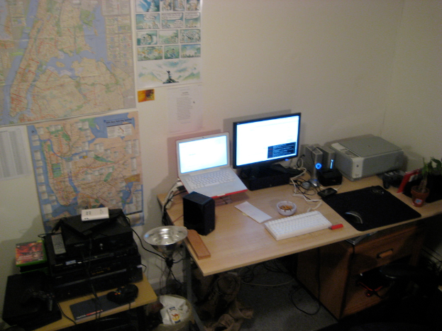 Ein Raum mit einem Tisch, auf dem ein Laptop, Lautsprecher, Monitor, Tastatur, Ball, Maus, Scanner und Papiere liegen, und einem weiteren Tisch mit Elektronikgeräten im Hintergrund, mit Charts und Postern an der Wand.