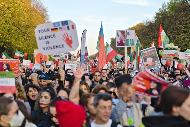 Eine große Menge hält Schilder und Flaggen mit Text, Bäume im Hintergrund unter einem klaren blauen Himmel, was auf eine Veranstaltung im Zusammenhang mit der iranischen Revolution hinweist.