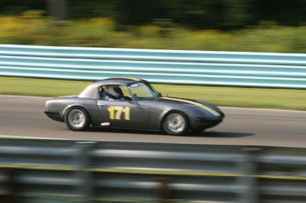 Ein Auto mit einem Helm tragenden Fahrer fährt eine Straße entlang, die von Barrieren gesäumt ist, mit Bäumen im Hintergrund.
