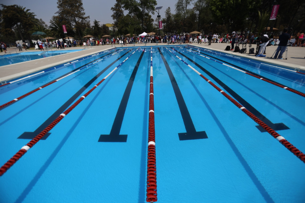 Großer Schwimmbad mit Bahnentrennung, umgeben von Menschen mit Taschen und Gegenständen, mit Zelten, Bannern, Stangen, einem Schild und einem bewölkten Himmel im Hintergrund.
