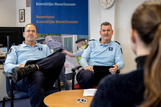 Zwei Polizeibeamte sitzen an einem Tisch mit einer Frau, Papieren und Gegenständen auf dem Tisch, einem Wandbanner und einer Uhr im Hintergrund, einem zweiten Tisch mit einem Monitor, Tastatur, Maus und Gegenständen links daneben und gerahmten Fotos an der Wand.