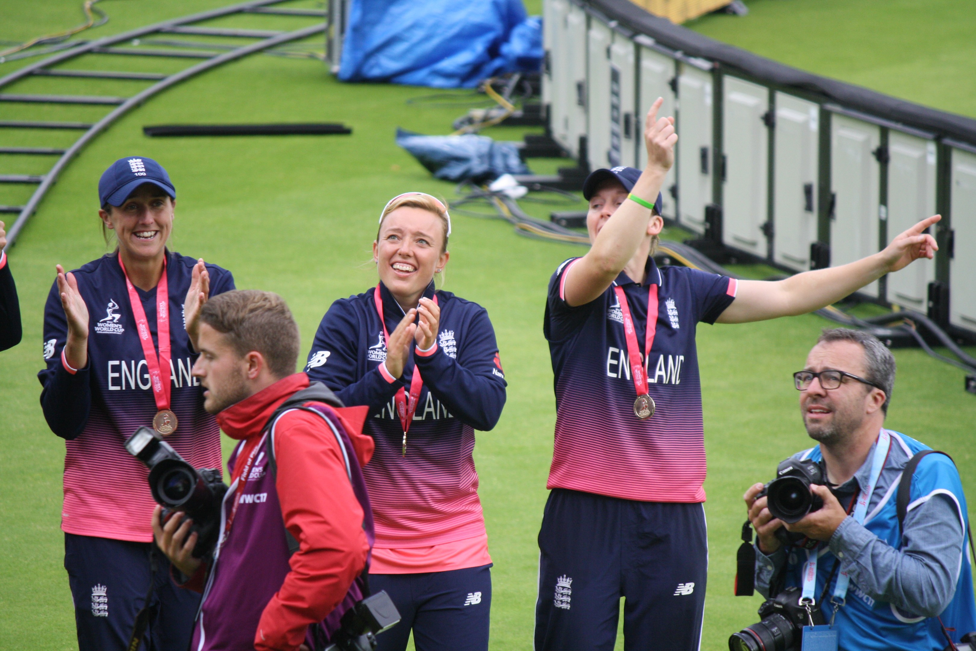 Gruppe von Menschen auf einem grünen Feld, die einen Sieg feiern, einige tragen Medaillen und Mützen, andere halten Kameras, mit Geländern und Taschen im Hintergrund.