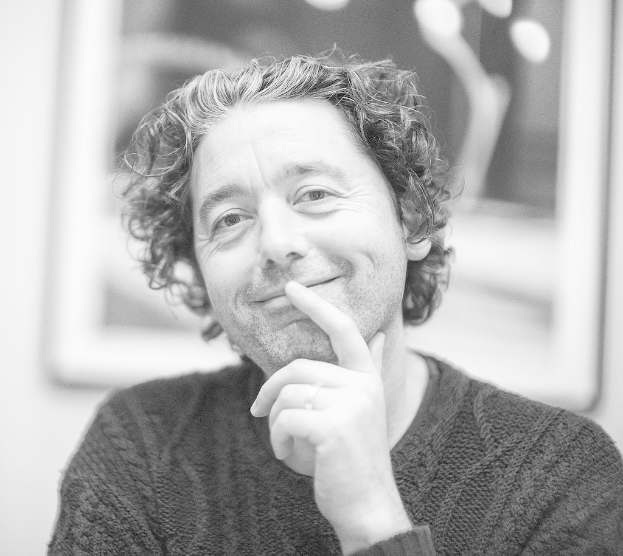 Ein Mann in einem Sweatshirt lächelt vor einem gerahmten Bild an der Wand.