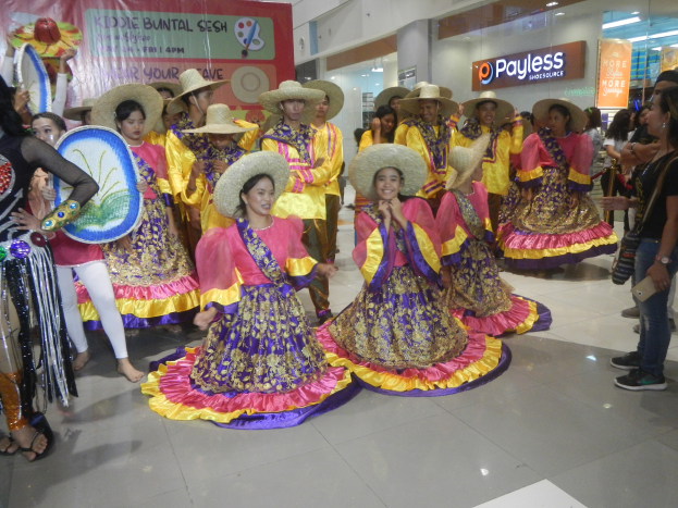 Eine Gruppe von Menschen in bunten Kleidern und Hüten tanzt in einem Einkaufszentrum, während andere in der Nähe stehen und Musikinstrumente halten, bei einer festlichen Cinco de Mayo-Feier.
