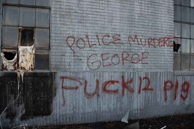 Ein Gebäude mit der Graffiti-Beschriftung "Polizei Mord George Fuck 12 P19" an der Seite, mit Fenstern und diversen Gegenständen vorne auf dem Boden.