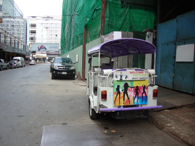 Gebäude, geparkte Autos, eine Werbetafel, ein bewölkter Himmel und ein Auto-Rikscha am Straßenrand.