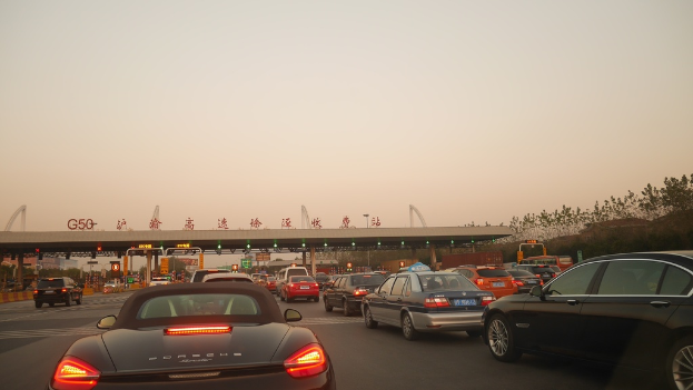 Fahrzeuge auf einer Straße mit einer Mautstation im Hintergrund, umgeben von Bäumen und Gebäuden, unter einem Himmel.
