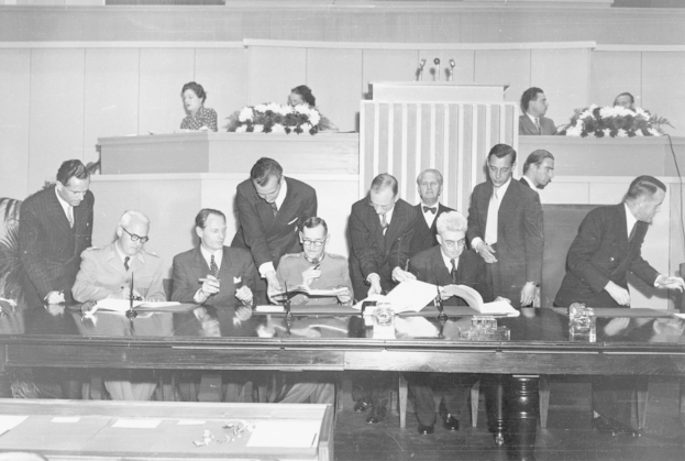 Eine Gruppe von Menschen, die auf Stühlen sitzen und andere dahinter an einem Tisch mit Mikrofonen, Gläsern und Blumensträußen.