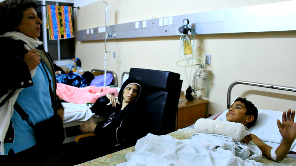 Ein Krankenhauszimmer mit mehreren Betten, Patienten, Infusionsflaschen auf Ständern und einer Frau mit einer Tasche in der Nähe eines Patienten.