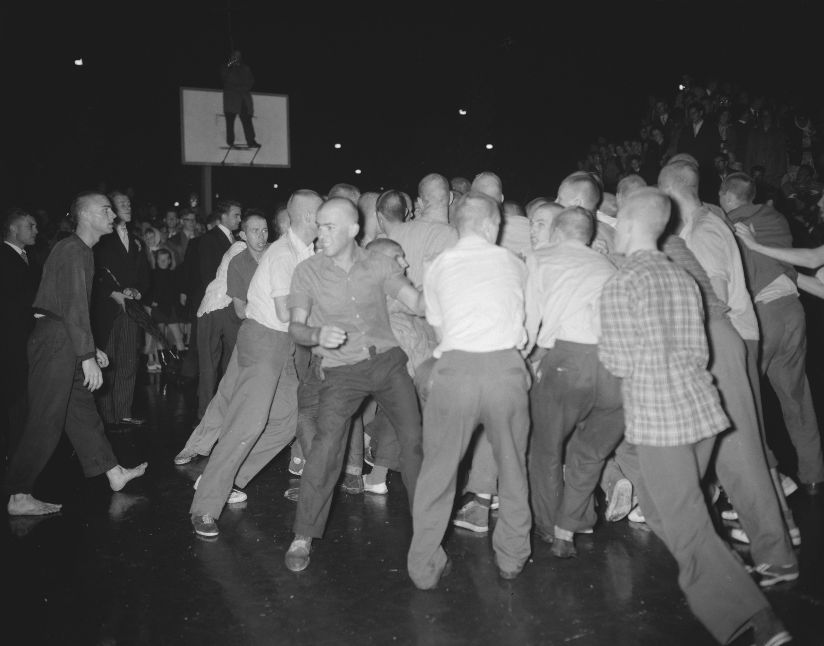 Black and white image of a group of men dancing on a stage with a screen and lights in the background.