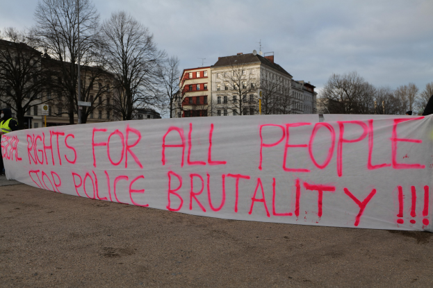 Group of people holding a banner reading "Rights for All People Stop Police Brutality" with a street pole, signboard, trees, buildings, and a cloudy sky in the background.