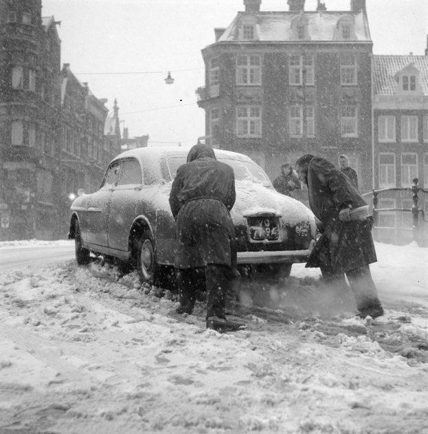 Menschen schippen Schnee von einem Auto auf einer verschneiten Straße, mit Gebäuden, einem Laternenmast und einem Himmel im Hintergrund.