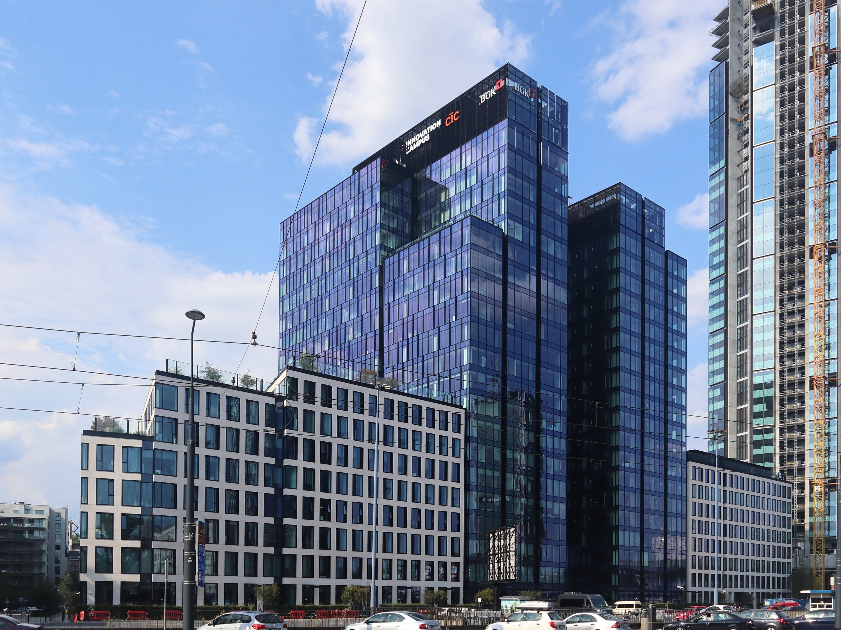 Großes modernes Bürogebäude mit vielen Fenstern, als Deutsche Bank Hauptsitz in Frankfurt identifiziert, umgeben von städtischer Infrastruktur einschließlich Straßenlaternen, Fahrzeugen, Bäumen und einem bewölkten Himmel.