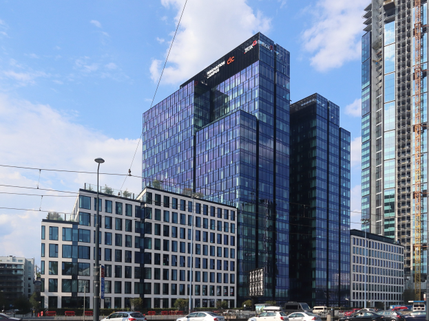 Großes modernes Bürogebäude mit vielen Fenstern, als Deutsche Bank Hauptsitz in Frankfurt identifiziert, umgeben von städtischer Infrastruktur einschließlich Straßenlaternen, Fahrzeugen, Bäumen und einem bewölkten Himmel.