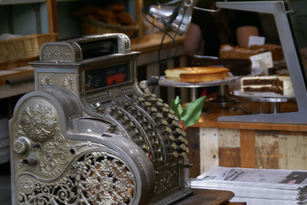Ein Tisch mit einer Maschine, Büchern, Kuchen auf Ständern, Papieren, einer Lampe und Körben aus Weide, die mit Gegenständen gefüllt sind.