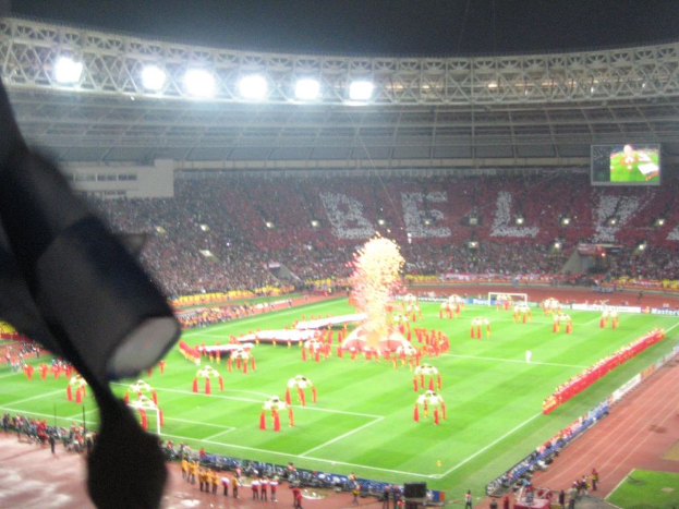 Ein Stadion mit Flutlicht, Eisengittern, Zuschauern, stehenden Individuen, Werbetafeln, Bildschirmen, Ãberwachungskameras, Fahnen und Deko.