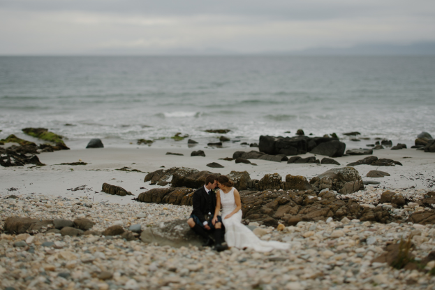 A bride and groom embrace and kiss on a rocky beach with the ocean and sky in the background.