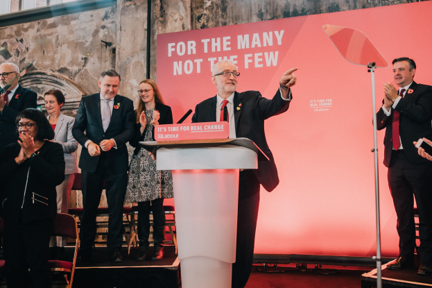 Ein Mann in einem schwarzen Blazer, weißem Hemd und roter Krawatte spricht in ein Mikrofon an einem Podium, mit einem Banner dahinter und einem Licht zu seiner Rechten, während eine Menge klatscht.