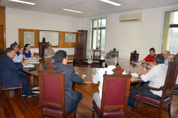 Eine Gruppe von Menschen, die an einem Tisch sitzen, mit Papieren und Gläsern, einem Klimagerät, einer Anschlagtafel und Karten an der Wand im Hintergrund.
