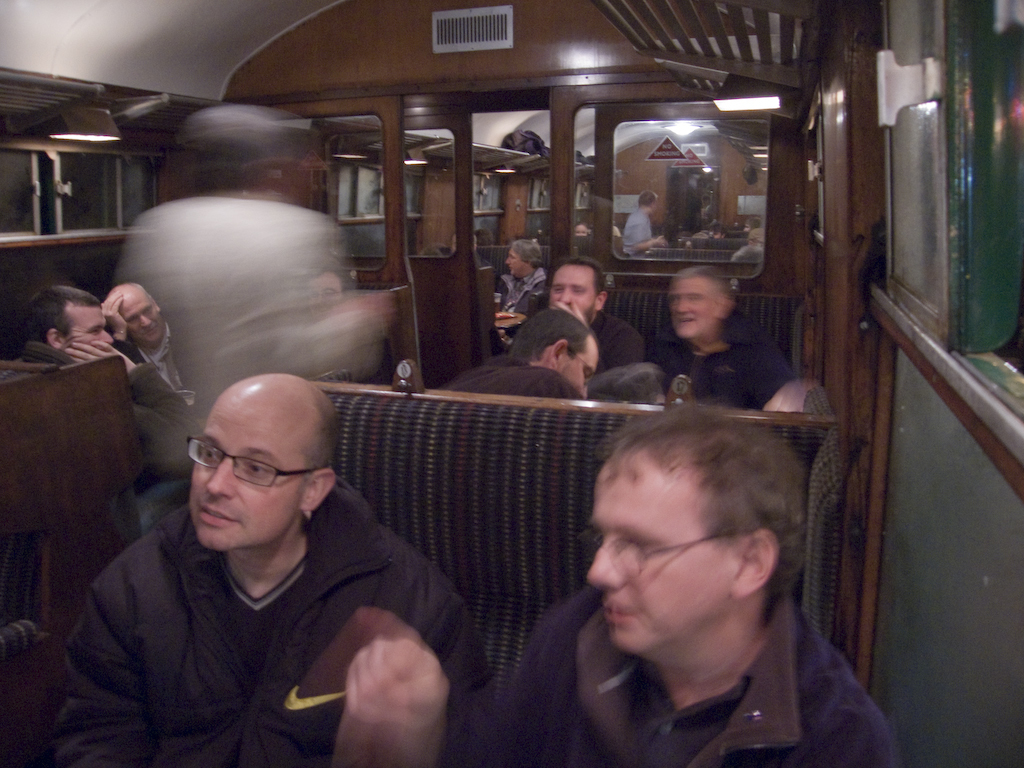 Zwei Männer sitzen in einem Zugabteil, hinter ihnen eine Gruppe von Menschen; oben ist ein Eingang zwischen den Abteilen zu sehen, rechts ein Fenster.