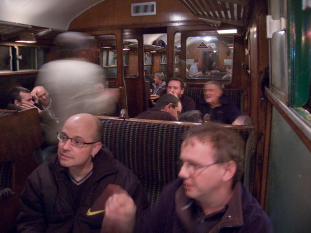 Zwei Männer sitzen in einem Zugabteil, hinter ihnen eine Gruppe von Menschen; oben ist ein Eingang zwischen den Abteilen zu sehen, rechts ein Fenster.