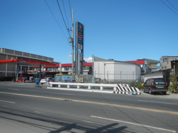 Tankstelle an einer Straßenecke mit parkenden Autos, umgeben von Gebäuden, Versorgungsmasten, Schildern, Geländern und Pflanzen unter einem bewölkten Himmel.