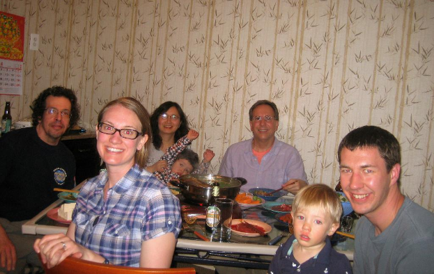 Ein Esstisch in einem Wohnzimmer mit verschiedenen Speisen, umgeben von fünf strahlenden Menschen, einem Kalender, einer Flasche und floralen Wanddesigns im Hintergrund und einem Stuhl im Vordergrund.