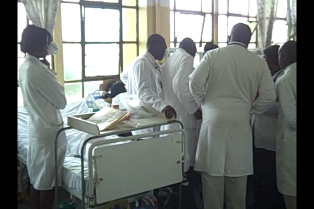 Arzte in einem Raum mit einem Bett und Gegenständen darauf, Vorhänge an Glasfenstern mit Metallstangen und durch die Fenster sichtbare Bäume.