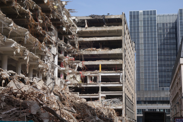 A large building in a city has been demolished, leaving debris surrounded by other buildings, street infrastructure, vehicles, trees, and a clear sky.