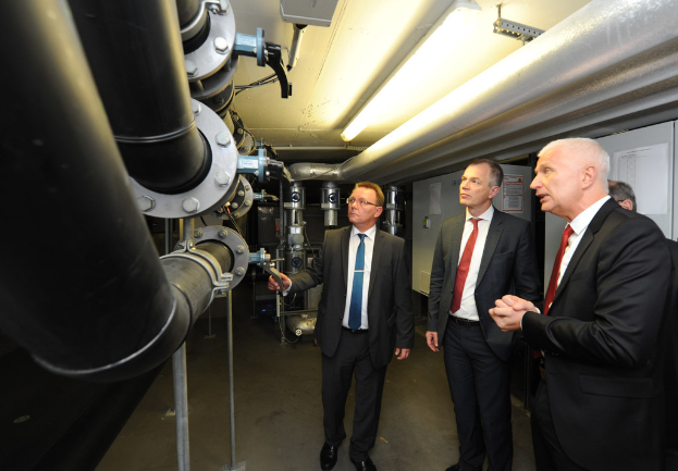 Vier Personen rechts, drei mit Mänteln, große Rohrleitungen links.