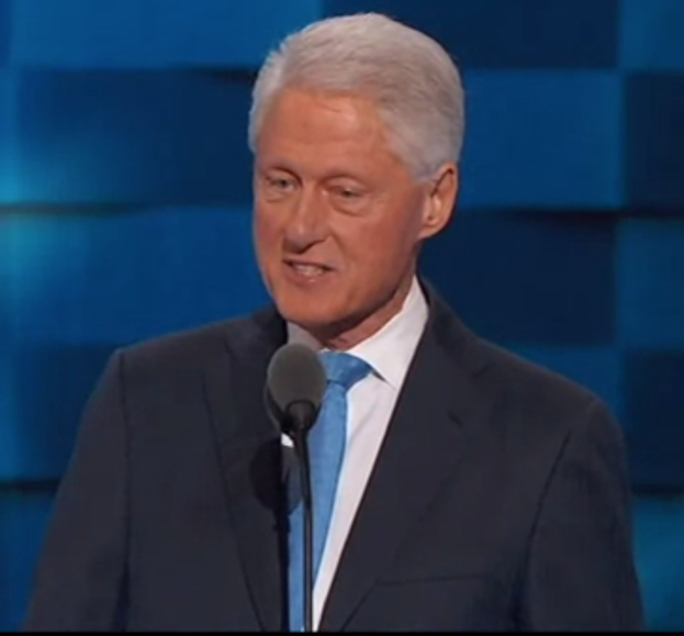 A man in a suit and tie stands at a podium with a microphone, giving a speech, with a wall in the background and a YouTube logo visible.