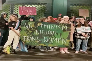 Eine Gruppe von Menschen hält ein Transparent mit der Aufschrift "Kein Feminismus ohne Trans-Frauen" und Schilder in einer öffentlichen Fläche mit einer Wand und einer Rolllade im Hintergrund.