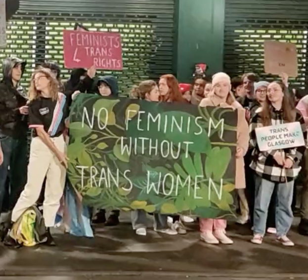 Eine Gruppe von Menschen hält ein Transparent mit der Aufschrift "Kein Feminismus ohne Trans-Frauen" und Schilder in einer öffentlichen Fläche mit einer Wand und einer Rolllade im Hintergrund.
