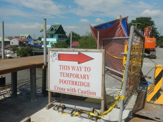 Temporäre Fußgängerbrücke mit Warnschild im Vordergrund, Gebäude und Bäume im Hintergrund und ein Fahrzeug auf der rechten Seite unter einem klaren blauen Himmel.