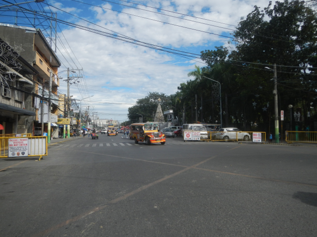 A busy city street with cars, buses, trucks, barricades, signboards, light poles, electric poles with wires, buildings with windows, trees, and a cloudy sky.