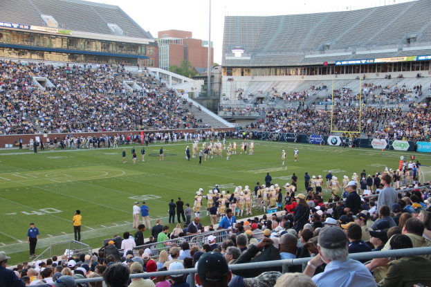 Ein Fußballspiel im Gange mit Spielern auf dem Feld und Zuschauern in den Rängen, in einem Stadion unter einem klaren blauen Himmel.