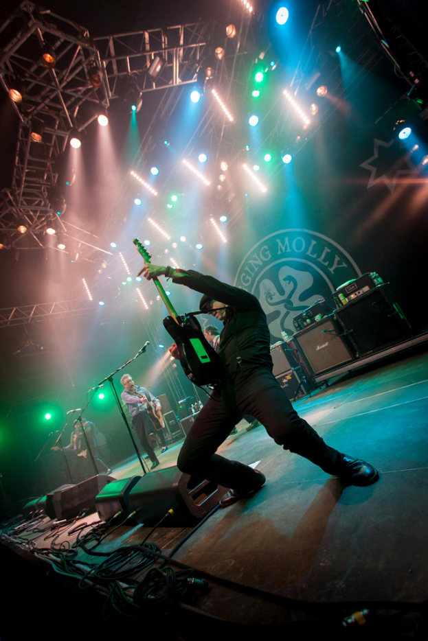 Ein musikalisches Konzert mit drei Personen auf der B├╝hne: zwei spielen auf der rechten Seite Gitarre und haben Mikrofone vor sich, ein Hintergrund mit 'Molly' und zwei Schlangenlogos sowie Scheinwerfer ├╝ber ihnen.