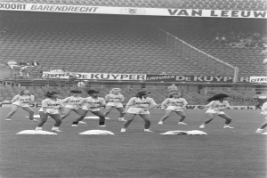 Eine Gruppe von Frauen auf einem Fußballfeld, umgeben von einer Menge auf den Tribünen und Schildern mit Text, in Schwarz-Weiß.