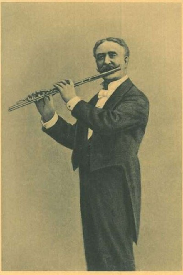 Old black-and-white photograph of a man in a tuxedo playing a flute on stage, eyes closed in concentration, with slicked-back hair and stage lighting illuminating his face.