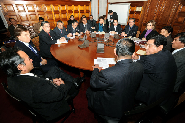Eine Runde Tischbesprechung mit Delegierten, Mikrofonen, Papieren, Kabeln und Dateien auf dem Tisch und einer hölzernen Wand im Hintergrund.